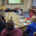 Szkolenie w bibliotece – prowadząca prezentuje zagadnienia na ekranie, uczestnicy siedzą przy stole i słuchają.