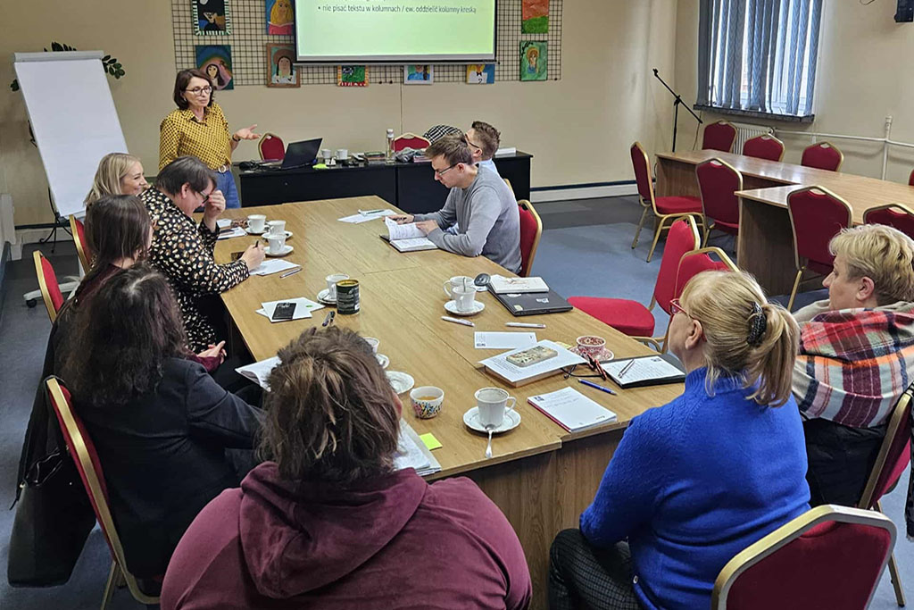 Szkolenie w bibliotece – prowadząca prezentuje zagadnienia na ekranie, uczestnicy siedzą przy stole i słuchają.