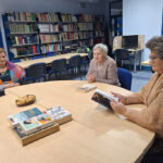 Trzy starsze kobiety siedzą przy okrągłym stole w bibliotece, rozmawiając o książkach. Na stole leżą egzemplarze książek oraz talerz z ciastem. W tle widać półki pełne książek oraz stanowiska komputerowe.