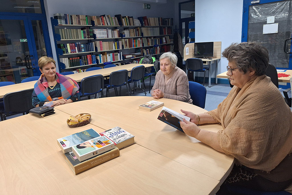 Trzy starsze kobiety siedzą przy okrągłym stole w bibliotece, rozmawiając o książkach. Na stole leżą egzemplarze książek oraz talerz z ciastem. W tle widać półki pełne książek oraz stanowiska komputerowe.