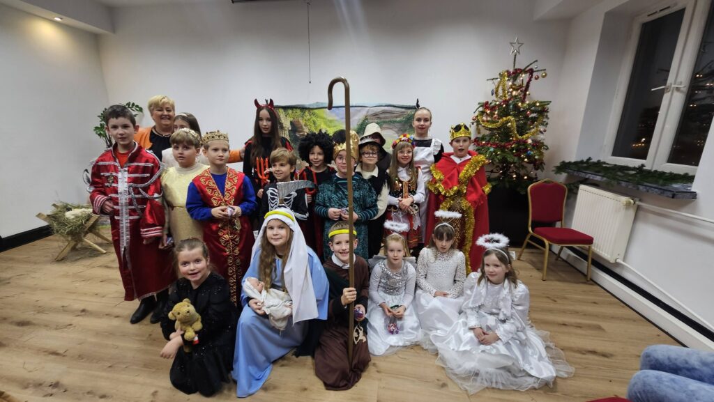 Grupa dzieci w świątecznych i jasełkowych strojach pozuje do wspólnego zdjęcia w sali, obok udekorowanej choinki; wśród kostiumów są anioły, królowie, pasterze i postacie biblijne.