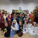 Grupa dzieci w świątecznych i jasełkowych strojach pozuje do wspólnego zdjęcia w sali, obok udekorowanej choinki; wśród kostiumów są anioły, królowie, pasterze i postacie biblijne