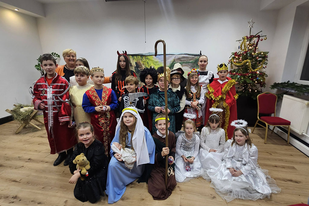 Grupa dzieci w świątecznych i jasełkowych strojach pozuje do wspólnego zdjęcia w sali, obok udekorowanej choinki; wśród kostiumów są anioły, królowie, pasterze i postacie biblijne